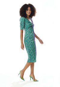 Green and purple patterned dress with a fitted silhouette, short puff sleeves, and a twisted front detail. Paired with green high-heeled shoes.
