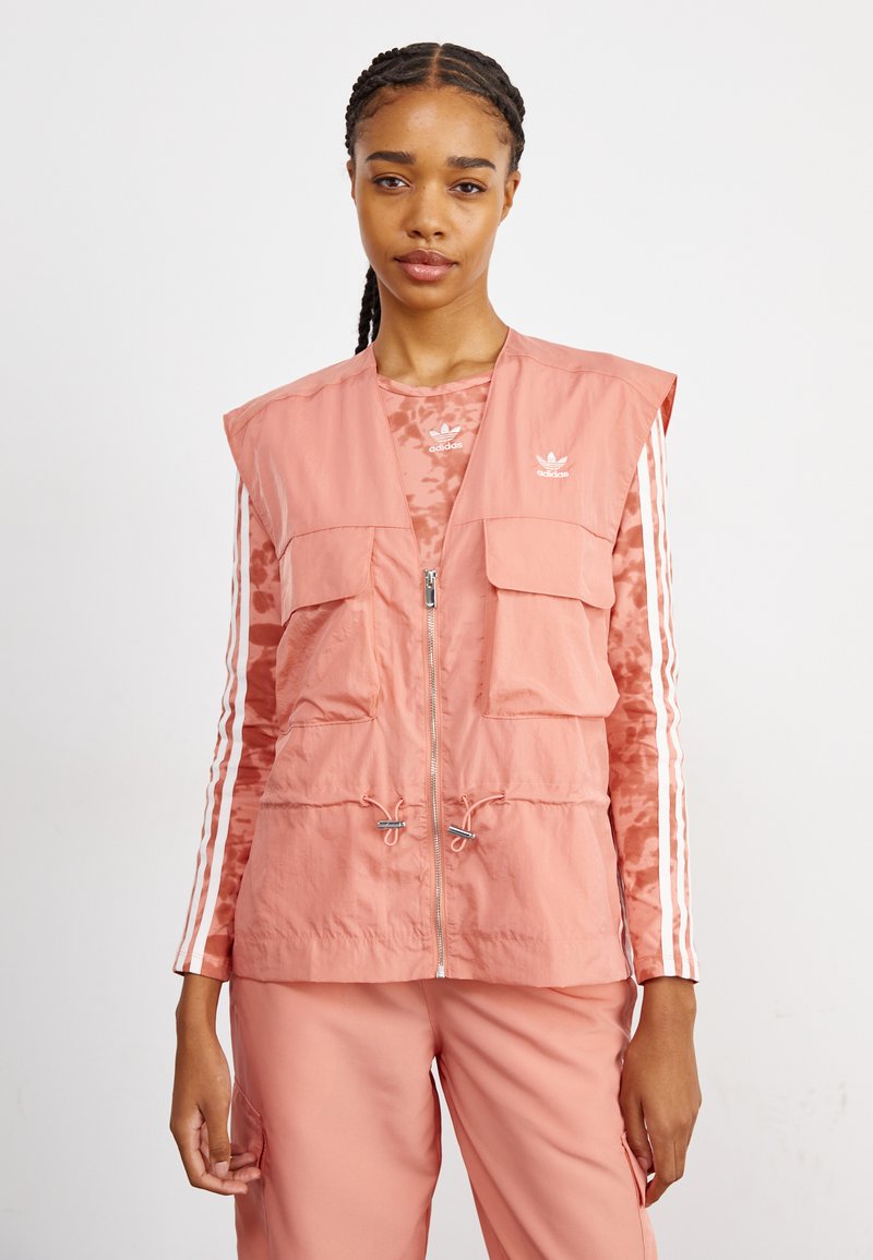 Ανοιχτό ροζ αμάνικο γιλέκο με φερμουάρ μπροστά, δύο μεγάλες τσέπες, κορδόνι στο κάτω μέρος και διακριτό λογότυπο. Συνδυάζεται με ένα μοντέρνο μπλούζα με μακρύ μανίκι και παντελόνι.