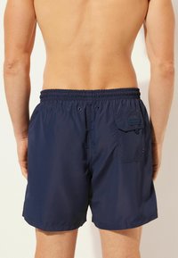 Marinblå badbyxor med elastisk midja, bakficka och förstärkta öglor; tillverkade av lätt, vattenavvisande tyg.