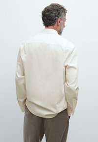 Camicia button-up a maniche lunghe color beige chiaro con una texture liscia, design con spalle a yoke sul retro e orlo arrotondato, abbinata a pantaloni grigi.