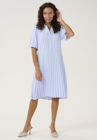 Svetlo modra plisirana obleka z V-izrezom in napihnjenimi rokavi, izdelana iz mehke, gladke tkanine in z aerodinamiko, v kompletu z bež natikači na peto.