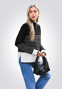 Schwarze und weiße gestreifte Strickpullover mit hohem Kragen, kombiniert mit blauen Denim-Jeans und einer schwarzen Lederhandtasche. Auffällige seitliche Schlitze.