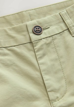 Ljusgröna bomullsshorts med en bronsknapp med texten "AUTHENTIC NEXT DUDE", synliga sömmar och en slät, matt yta.