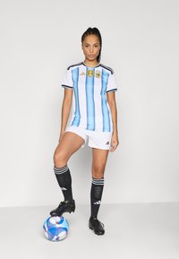 Kvinna står med ena foten på en blåvit fotboll, bär Argentinas fotbollströja, vita shorts, svarta strumpor och fotbollsskor mot en enfärgad bakgrund.
