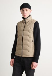 Beige dunjakke med vertikale quiltede seksjoner og høy krage, paret med en svart strikket genser, satt mot en nøytral bakgrunn.