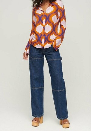 Femme portant une chemise à manches longues colorée et à motifs, un jean bleu foncé à larges jambes et des sandales compensées en bois couleur beige, debout sur un fond uni.