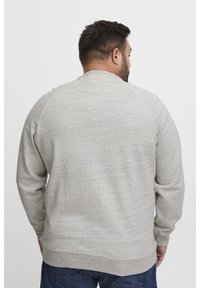 Man met kort donker haar in een lichtgrijze lange mouwen trui en donkerblauwe jeans, met de achterkant naar de camera gericht tegen een plain witte achtergrond.