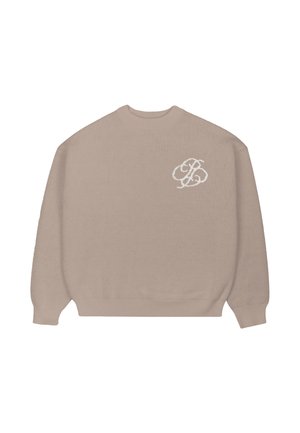 Mężczyzna ubrany w beżowy sweter z białym logo, brązowe spodnie w prążki i kremową zimową czapkę trzyma zieloną kurtkę, patrzy w bok.