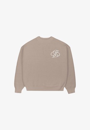 Mężczyzna ubrany w beżowy sweter z białym logo, brązowe spodnie w prążki i kremową zimową czapkę trzyma zieloną kurtkę, patrzy w bok.