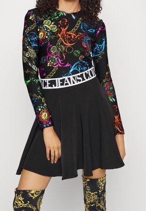 Femme portant une robe noire avec un haut à motif baroque coloré, une ceinture blanche avec texte, et des bottes montant jusqu'aux genoux assorties au motif.