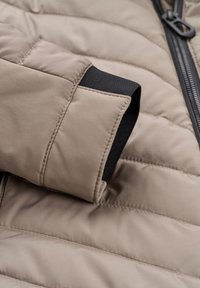 Beige Pufferjacke mit gesteppte Abschnitten und schwarzem elastischen Bündchen. Verfügt über einen Reißverschluss und glattes, wasserabweisendes Material.