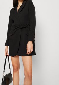 El vestido blazer negro con cinturón presenta un ajuste entallado, un diseño de envoltura, mangas largas con acentos de botones y una tela de textura suave.
