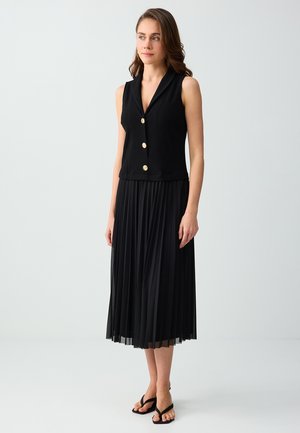 Kobieta w czarnej bezrękawnikowej kamizelce na guziki, czarnej plisowanej spódnicy midi i czarnych sandałach, stojąca na jednolitym jasnym tle.