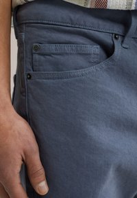 Modré bavlnené nohavice majú predné vrecko s gombíkom a špeciálnym prešívaním. Tkanina má hladkú textúru s jemným leskom.