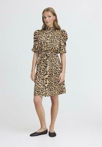 Robe en imprimé léopard avec des manches bouffantes, une fermeture à boutons à l'avant et une taille nouée. Présente un col et un ourlet au niveau des genoux. Portée avec des ballerines noires.