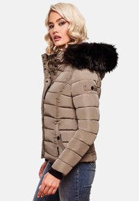 Puffjakke i taupe med hætte af sort faux fur. Har kviltede sektioner, lynlås og trykknapper. Bæres med blå jeans.