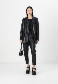 Zwarte faux leren blazer met een reverskraag, gecombineerd met bijpassende slim fit broek en chunky platform schoenen. Het model houdt een kleine handtas vast.