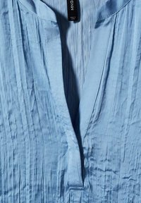 Camicia azzurra chiara realizzata in tessuto testurizzato, con pieghe verticali, scollatura a V e un'etichetta che recita "MNG."