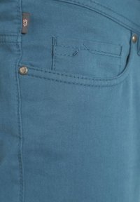 Hellblaue Denim-Stoff mit Textur, versehen mit einer kleinen Fronttasche und einem metallischen Knopfakzent. Minimale Nahtdetails sind sichtbar.