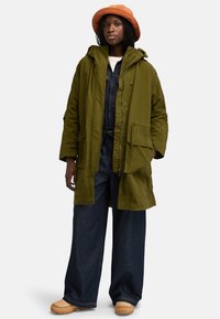 Olivgrön parkas med huva, flera fickor och dragkedja. Bärs över mörka denimsoveraller och matchas med beige träskor och en persikofärgad hatt.