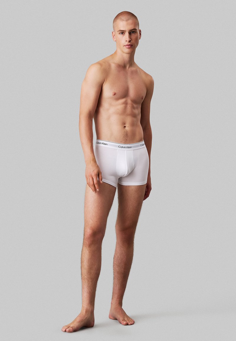 Hvide bomulds boxershorts med en blød elastisk talje, der har Calvin Klein-logoet. Modellen står barfodet og viser et tætsiddende design.