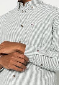 Camisa de botones a rayas en verde y blanco, con un bolsillo en el pecho, botones marrones y una pequeña etiqueta con logo en el lado izquierdo.