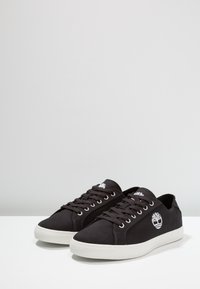 Baskets en toile noire avec semelles en caoutchouc blanc, dotées d'œillets argentés et d'un logo d'arbre blanc sur le côté. Bout arrondi et design à lacets.