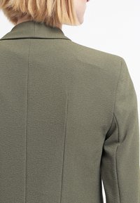 Olivově zelený blazer se strukturovaným zadním dílem, texturovanou tkaninou a jemnými detaily švů. Klasický klopový límec a přiléhavý střih.