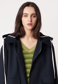 Marineblaues Kapuzenshirt mit cremefarbenen Kordeln und Fronttaschen, über einem grünen gemusterten Strickoberteil getragen. Glatte Stoffstruktur.