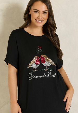 Femme souriante portant un t-shirt noir avec deux oiseaux en sequins qui se regardent sous le gui et le texte "Bisous de Noël" à l'avant.