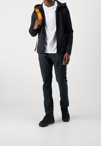 Veste noire à capuche portée sur un t-shirt blanc, associée à un pantalon sombre et des chaussures noires. La veste comporte une sangle orange sur une épaule.