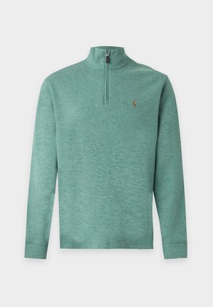 Homme portant un pull vert à fermeture éclair au quart, un pantalon cargo beige et des baskets blanches, debout devant un fond clair uni.