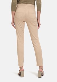 Personne portant un pantalon slim beige et des sandales à talons blocs beiges, vue de dos sur un fond blanc uni.