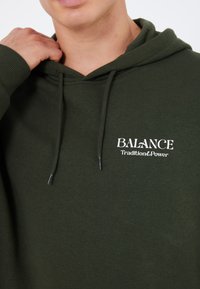 Hoodie vert olive en tissu doux, avec une poche kangourou à l'avant, des cordons de serrage, et un texte brodé blanc "ÉQUILIBRE Tradition&Puissance."
