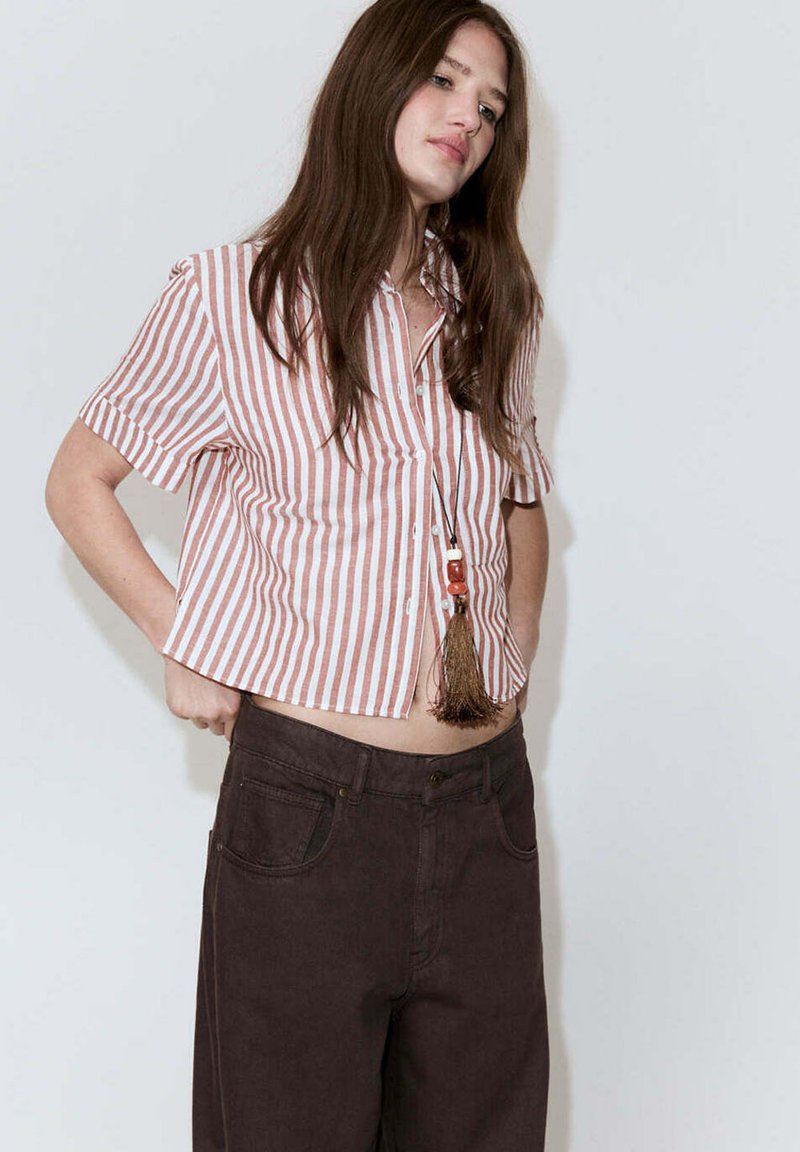 Jeune femme ajustant un pantalon marron foncé, portant une chemise à manches courtes rayée rouge et blanche à boutons et un collier à pompons.