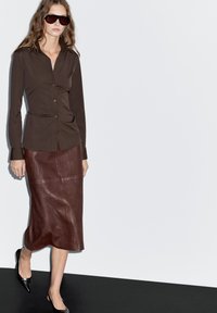 Bruine button-up shirt met slanke lijnen, gecombineerd met een knielange bordeaux leren rok en puntige zwarte flats. De outfit heeft een minimalistisch ontwerp.