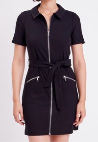 Robe noire à manches courtes avec fermeture éclair frontale, ceinture nouée à la taille et deux poches zippées, portée par une personne debout sur fond blanc.