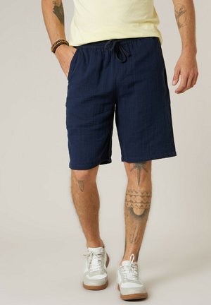 Homme portant un short bleu marine à cordon, des baskets blanches et un t-shirt jaune, montrant des tatouages sur les jambes et les bras.