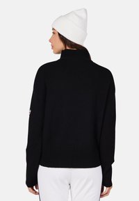 Maglione nero lavorato a maglia con collo alto e vestibilità ampia, con polsini e orlo a costine; abbinato a un berretto bianco.