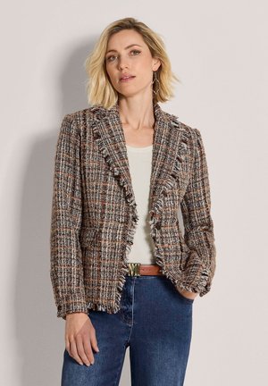 Frau mit blondem Haar trägt einen braunen karierten strukturierten Blazer über einem weißen Top und blauen Jeans und steht vor einer schlichten hellen Wand.