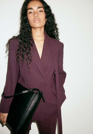 Young woman with long curly hair wearing a plum-colored suit holding a black leather handbag against a plain light background.