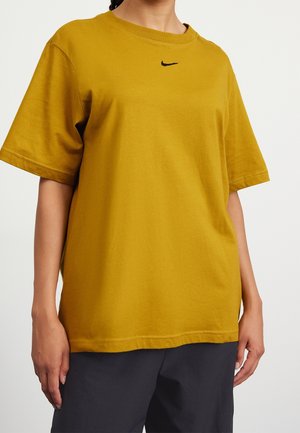 Persoon draagt een mosterdgele korte mouwen Nike T-shirt en donkere broek, staat voor een effen lichte achtergrond.