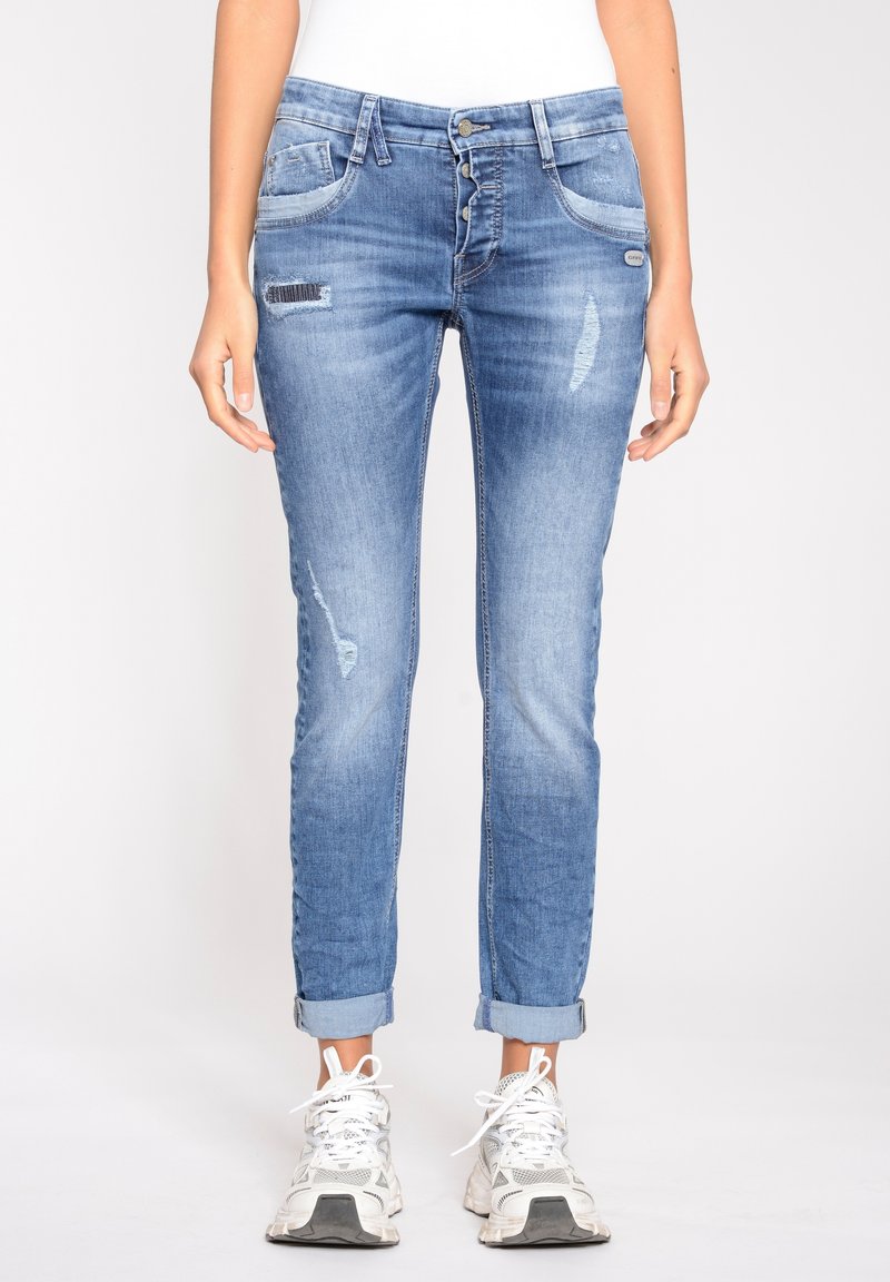 Personne portant un jean bleu déchiré retroussé et des baskets blanches, debout devant un fond clair uni.