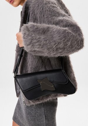 Sac en cuir noir avec un accent en argent ondulé, porté par une personne dans un manteau gris en peluche. Présente un design épuré et une bandoulière réglable.