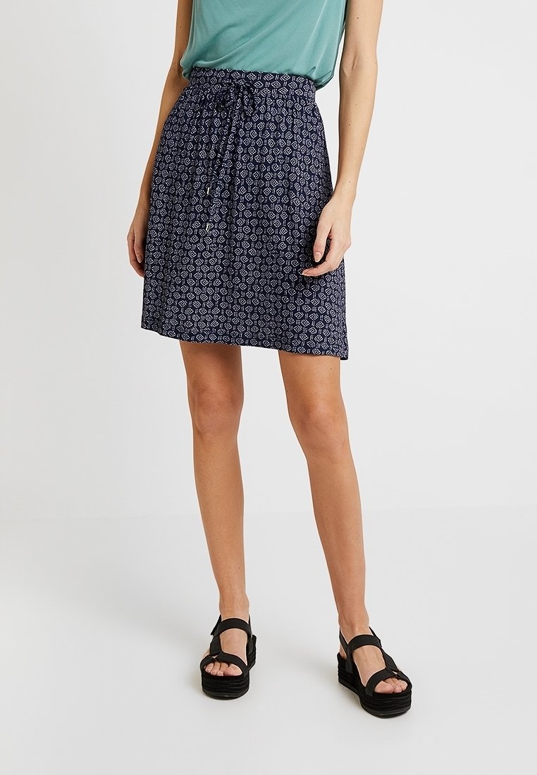 Jupe bleu marine à motifs avec taille à cordon, longueur genou, ornée de dessins géométriques. Associée à des sandales à plateforme noires.