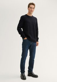 Donkerblauwe gebreide trui met een textuurpatroon, ronde hals. Gecombineerd met blauwe jeans en zwarte enkellaarsjes, staand tegen een lichte achtergrond.