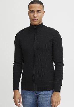 Svart teksturert zip-up genser med ribbet krage og mansjetter, med en glatt front og en tettsittende design. Båret over blå jeans.