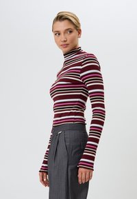 Langarm turtleneck top met horizontale strepen in bordeaux, roze, crème en blauw. De stof lijkt ribbelig en gestructureerd. Gecombineerd met grijze broek.
