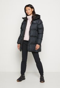 Moteris stovi juodo pūkinio švarko, vilkėdama šviesiai rožinės spalvos megztinį, juodas kelnes ir juodas sportinius batelius prieš baltą foną.