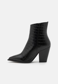 Botte de cheville noire avec un bout pointu, en cuir verni embossé crocodile, présentant un talon épais et une texture lisse et brillante.